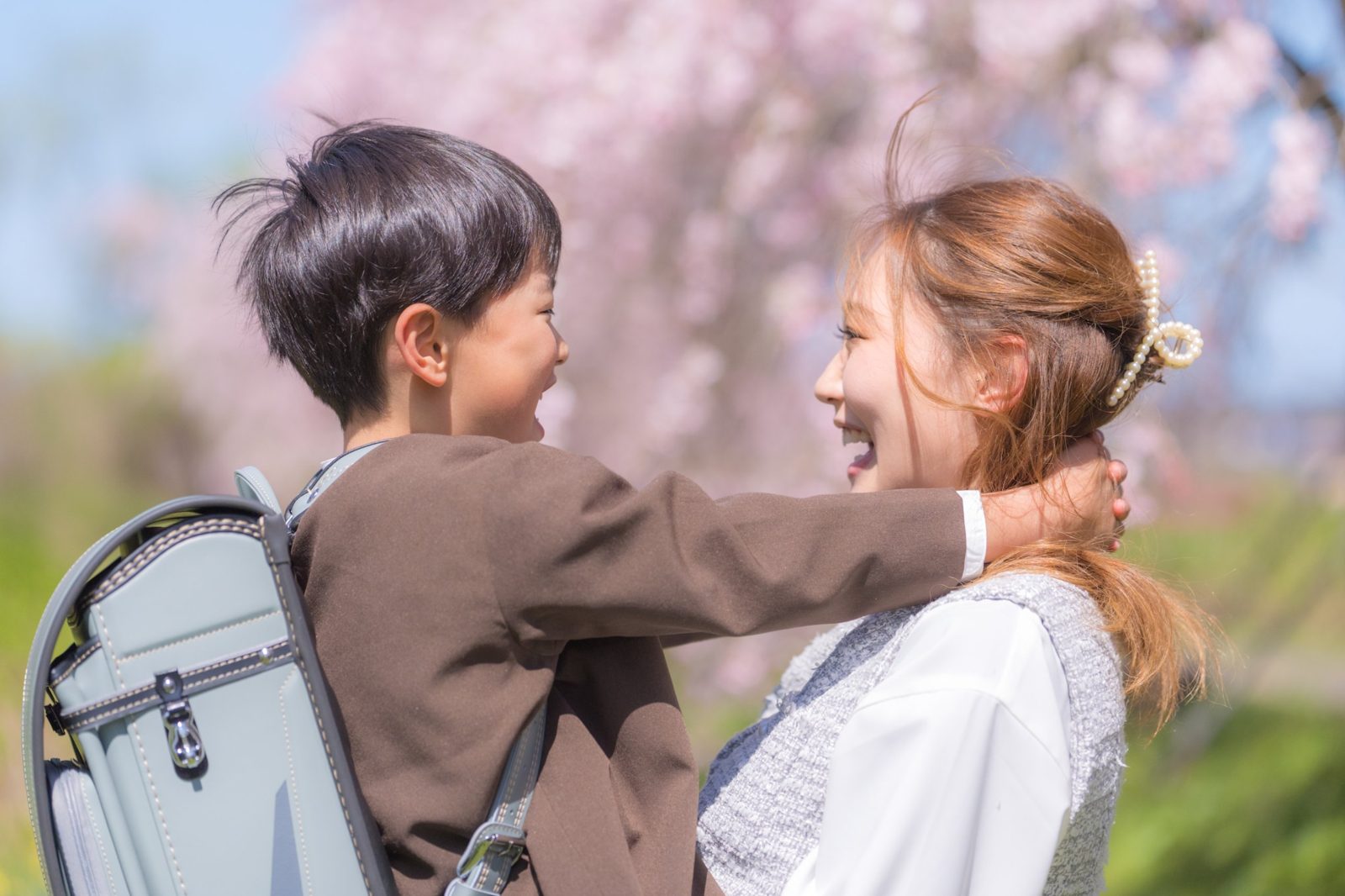 桜の下で、ランドセルを背負った男の子がお母さんに抱きついて笑う